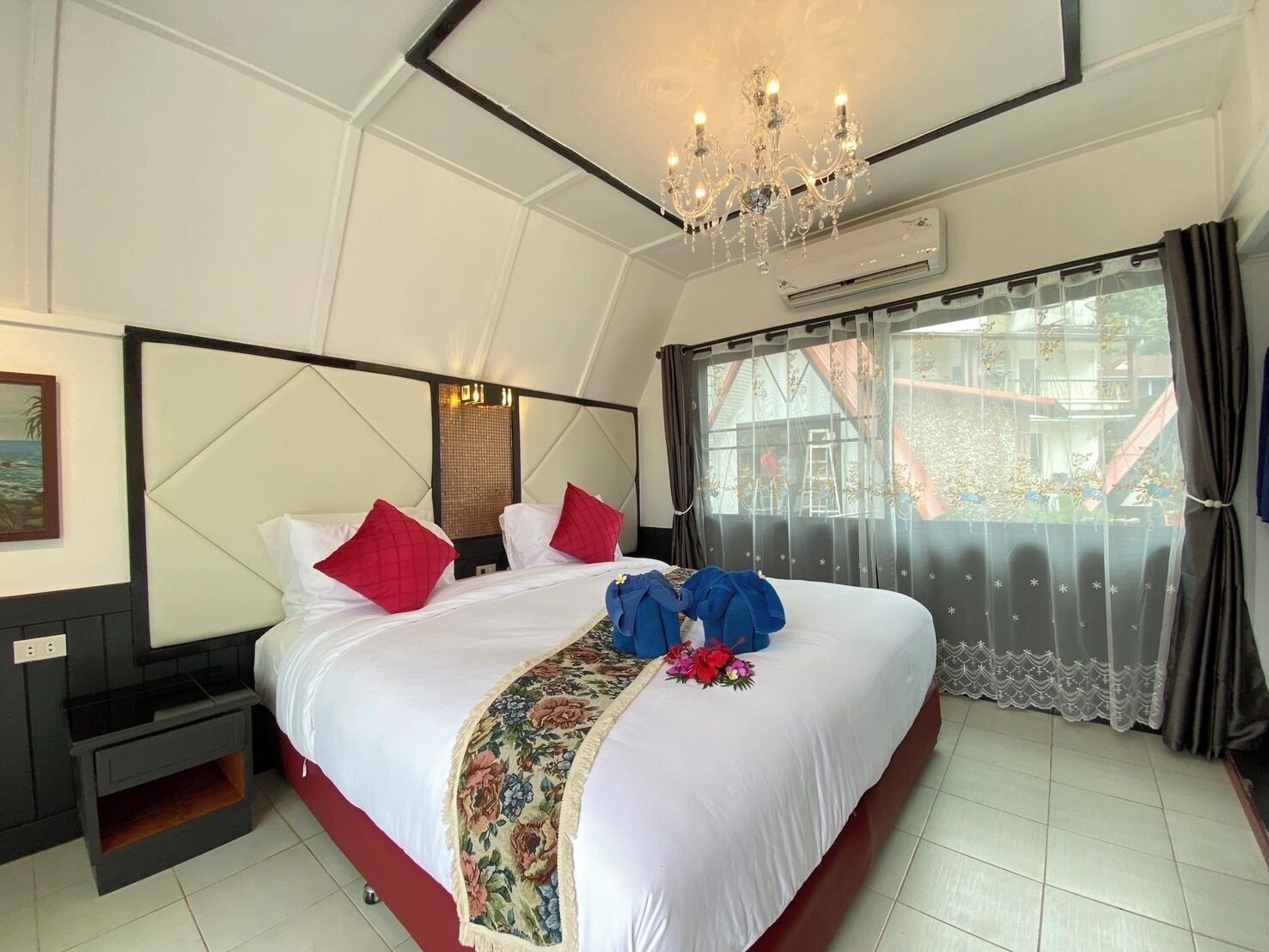 A hotel room with a large bed, a chandelier, a painting on the wall, and a window with curtains.