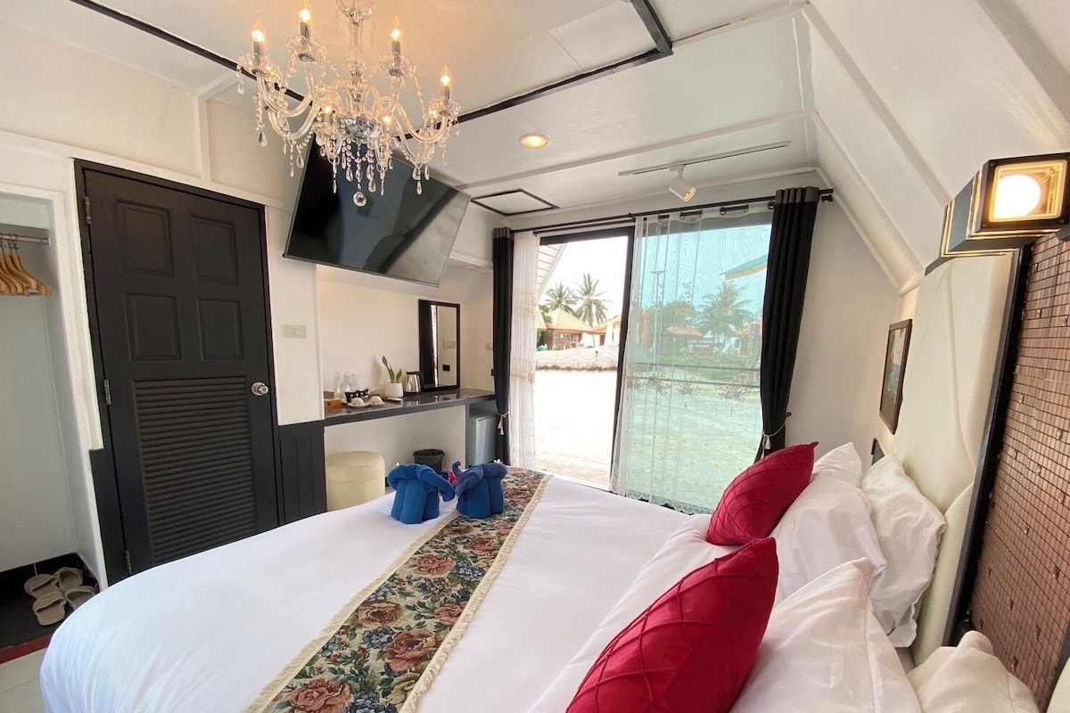 A hotel room with a large bed, a chandelier, a view through a sliding door, and a small kitchenette.