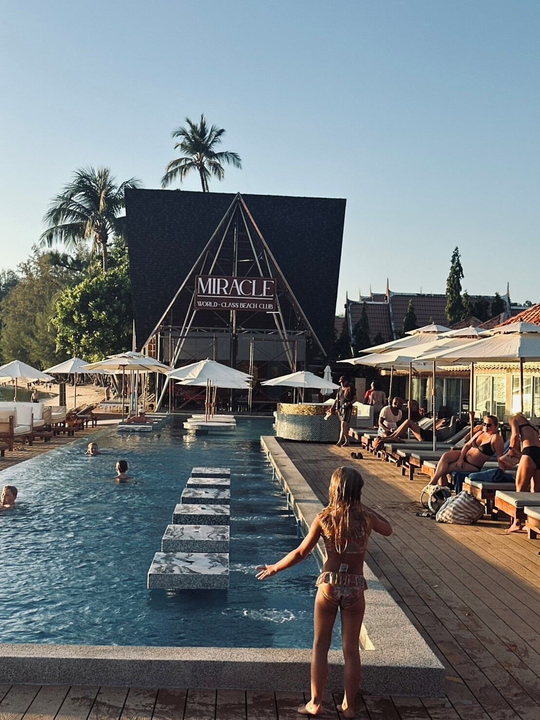 A pool area with a large sign reading "Miracle World-Class Beach Club" and a wooden deck with lounge chairs and umbrellas.