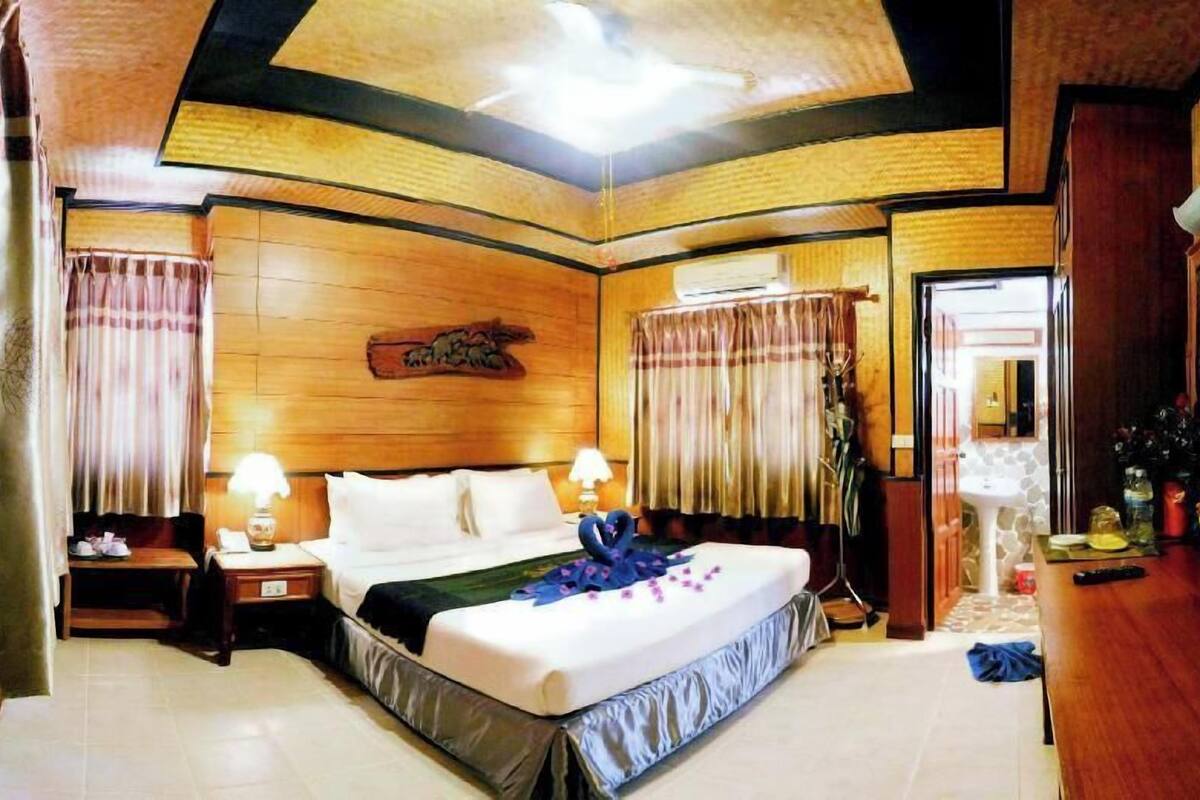 A hotel room with a bed, two bedside tables, a nightstand, a wall-mounted air conditioning unit, a bathroom with a sink, and a mirror.