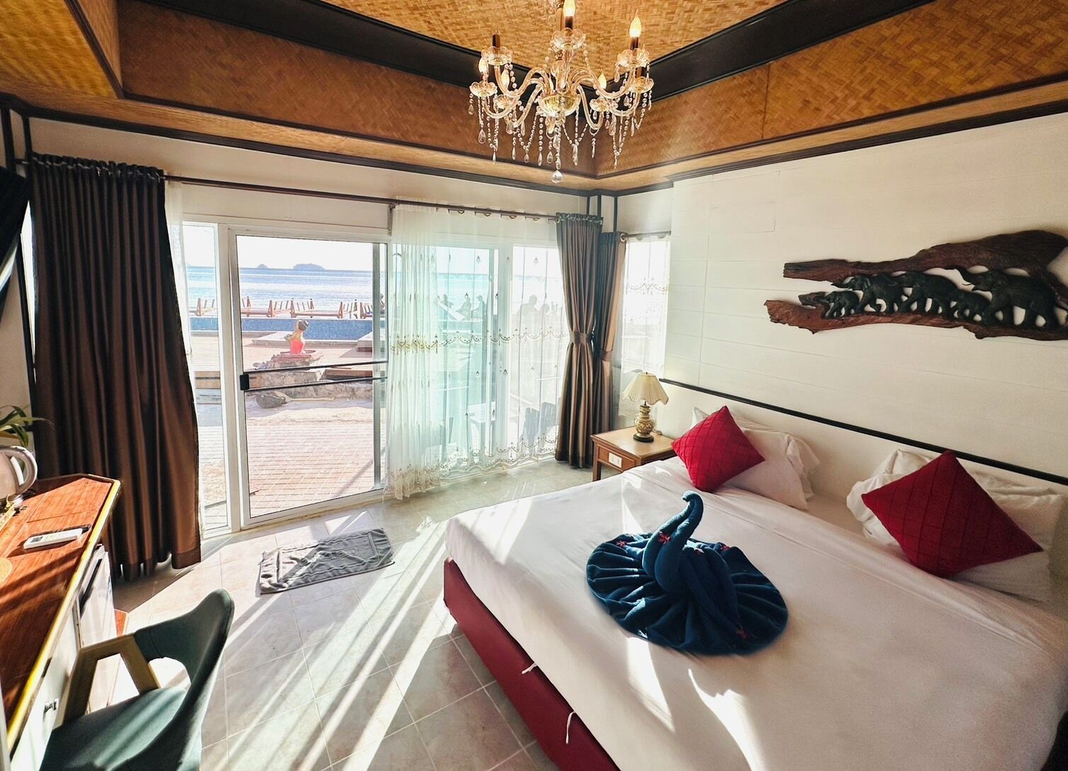 Een hotelkamer met een bed, een bureau, een stoel, een kroonluchter en uitzicht op het strand.