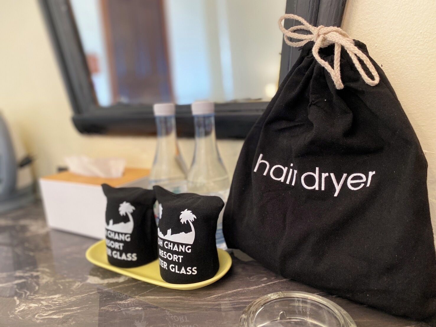 Una bolsa negra con cordón de ajuste y la leyenda "haiji dryer", dos calcetines negros con la inscripción "DH CHANG RESORT WATER GLASS" y una botella de agua de vidrio transparente sobre una mesa.