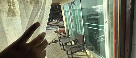 Una mano toca una cortina translúcida, mostrando un balcón con suelo de baldosas y puerta de cristal.