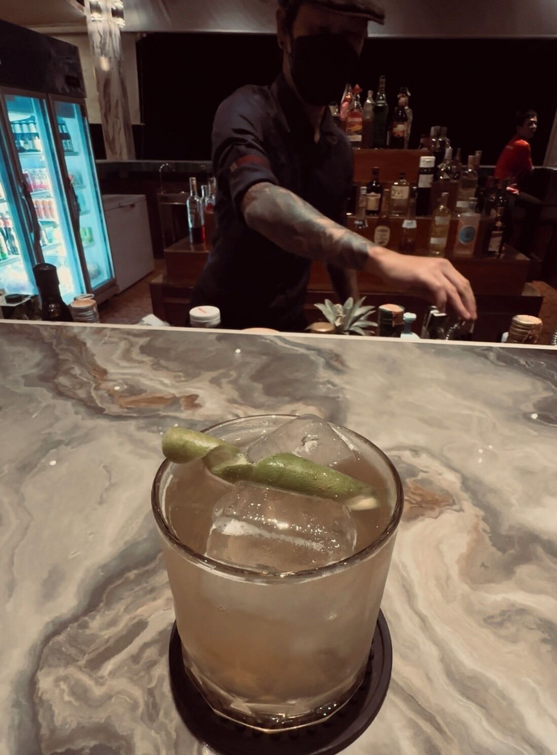 A bar area with a marble countertop, a glass with a green garnish, and a refrigerator in the background.