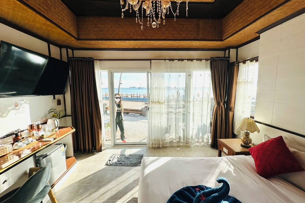 A hotel room with a bed, a desk with a TV, a chandelier, and a view of the beach through the window.
