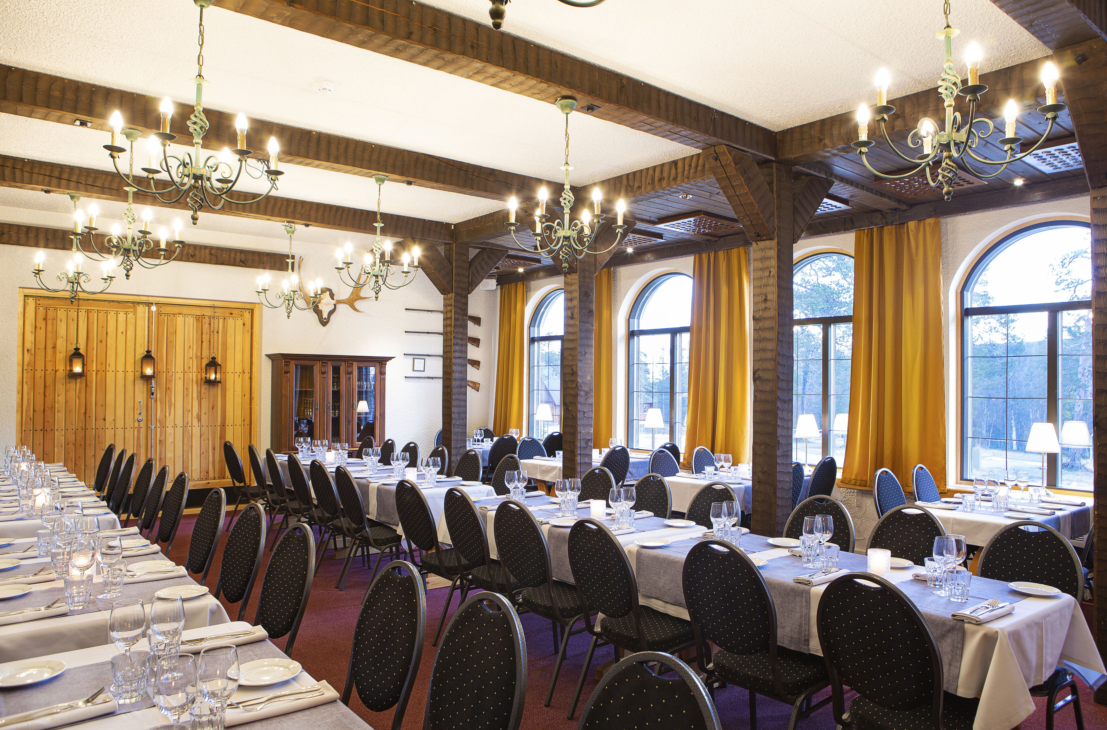 A banquet hall with long tables set for dining, chandeliers, and large windows with curtains.