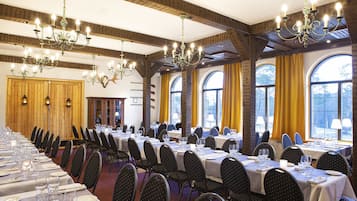 A banquet hall with long tables set for dining, chandeliers, and large windows with curtains.