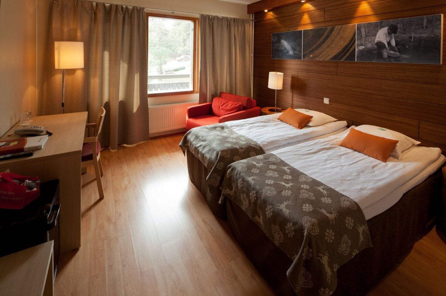 A hotel room with a wooden floor, a desk with a lamp, a red chair, a bed with two pillows, and a window with curtains.