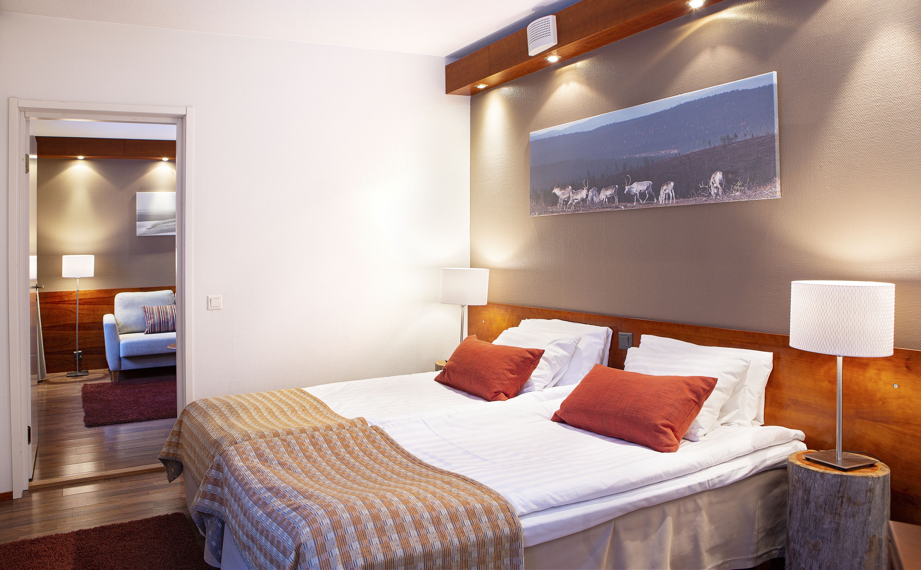 A hotel room with a large bed, a wooden headboard, a painting of a herd of deer, and a nightstand with a lamp.