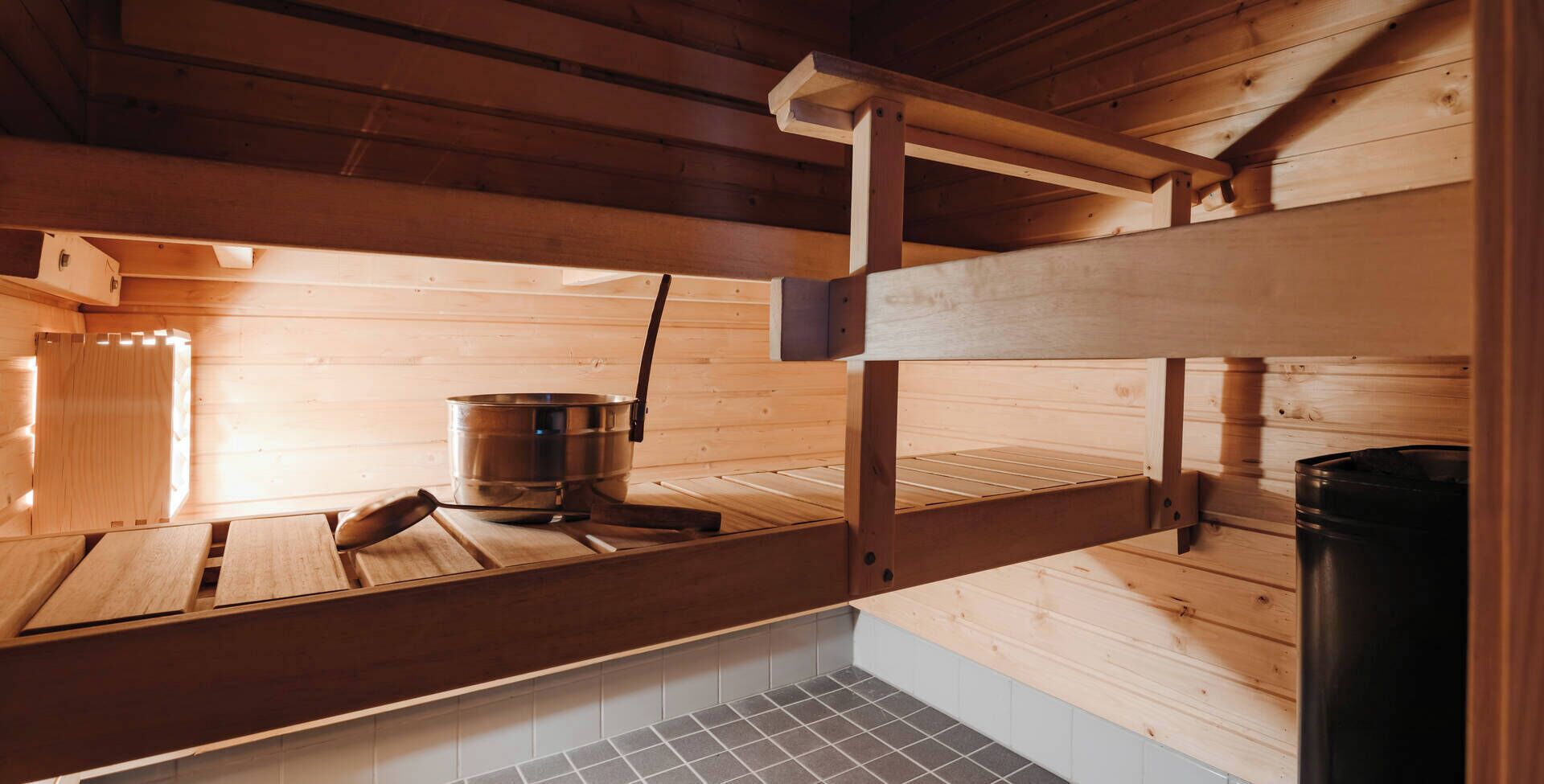Una sauna de madera con una olla de metal, un banco de madera y piso de baldosas.