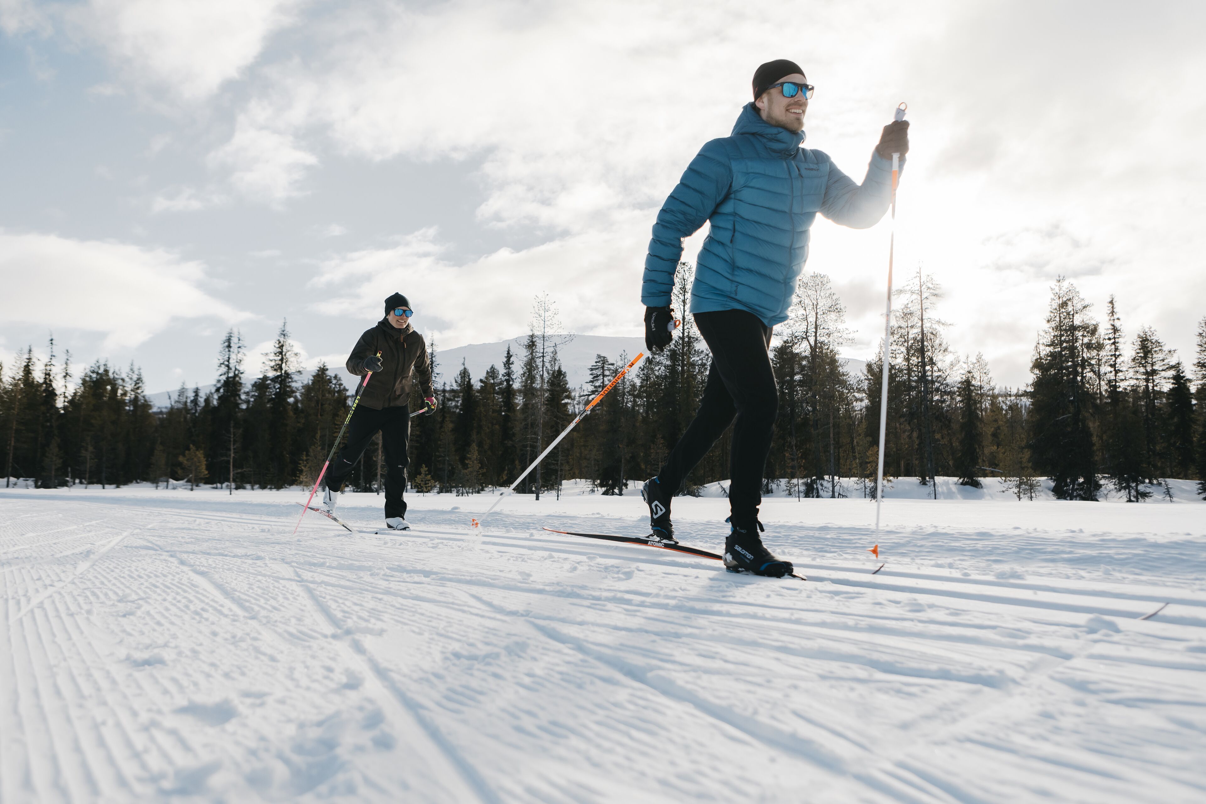 Two people cross-country skiing on a snowy trail with trees and mountains in the background.