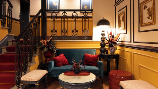 A room with a staircase, a blue sofa with red cushions, a round table with a tray, and a red ottoman. - JOIVY Together Florence Secret Nest (Florence)