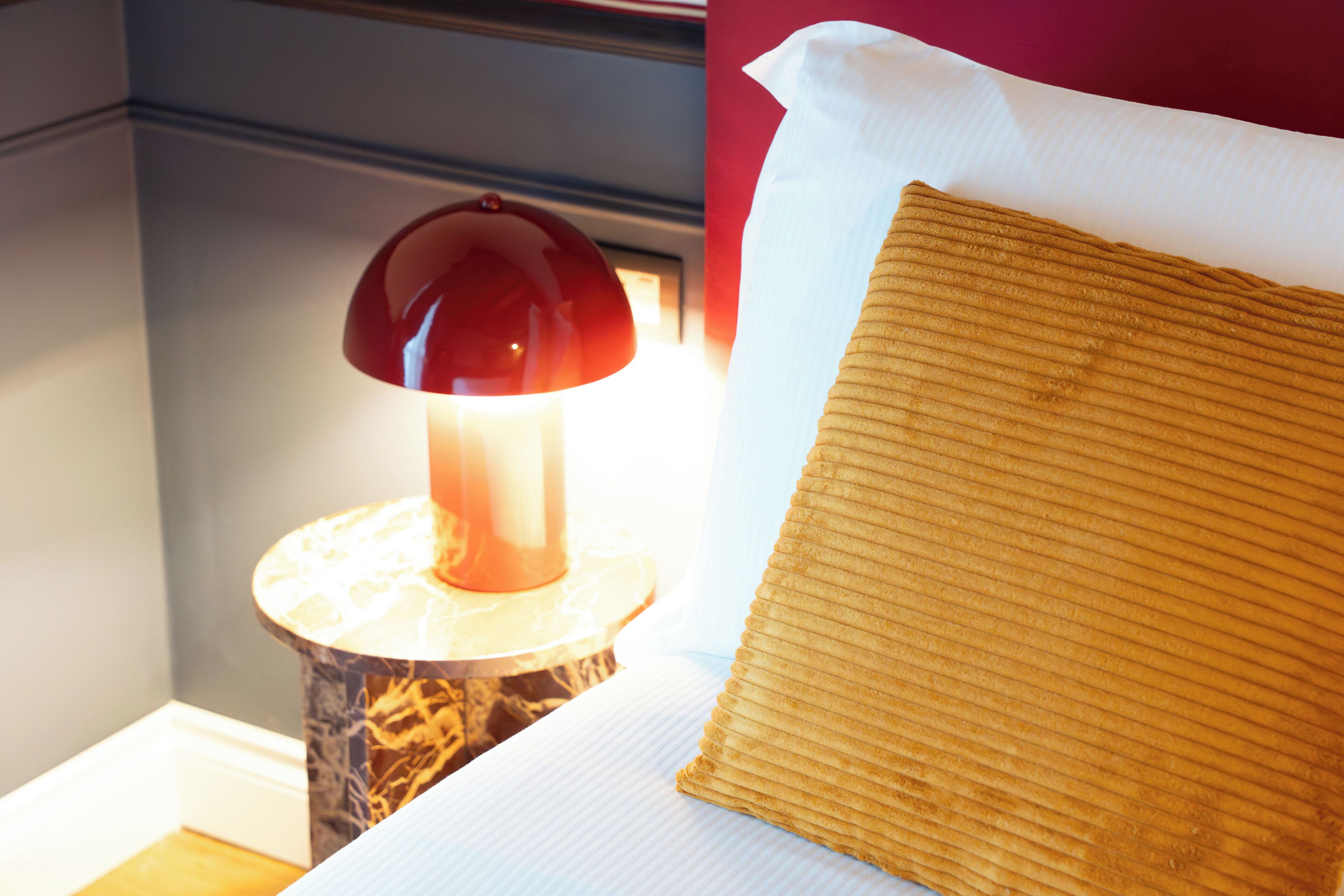A neatly made bed with a white pillow and a textured brown pillow, a red lamp on a marble base, and a wall with a red accent.