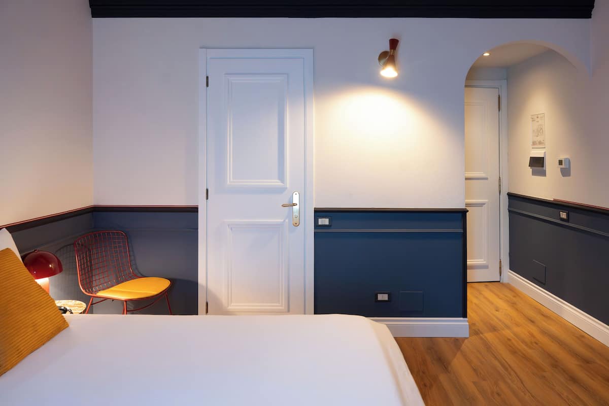 A hotel room with a bed, a chair, a desk, a door, and a wall-mounted light fixture.