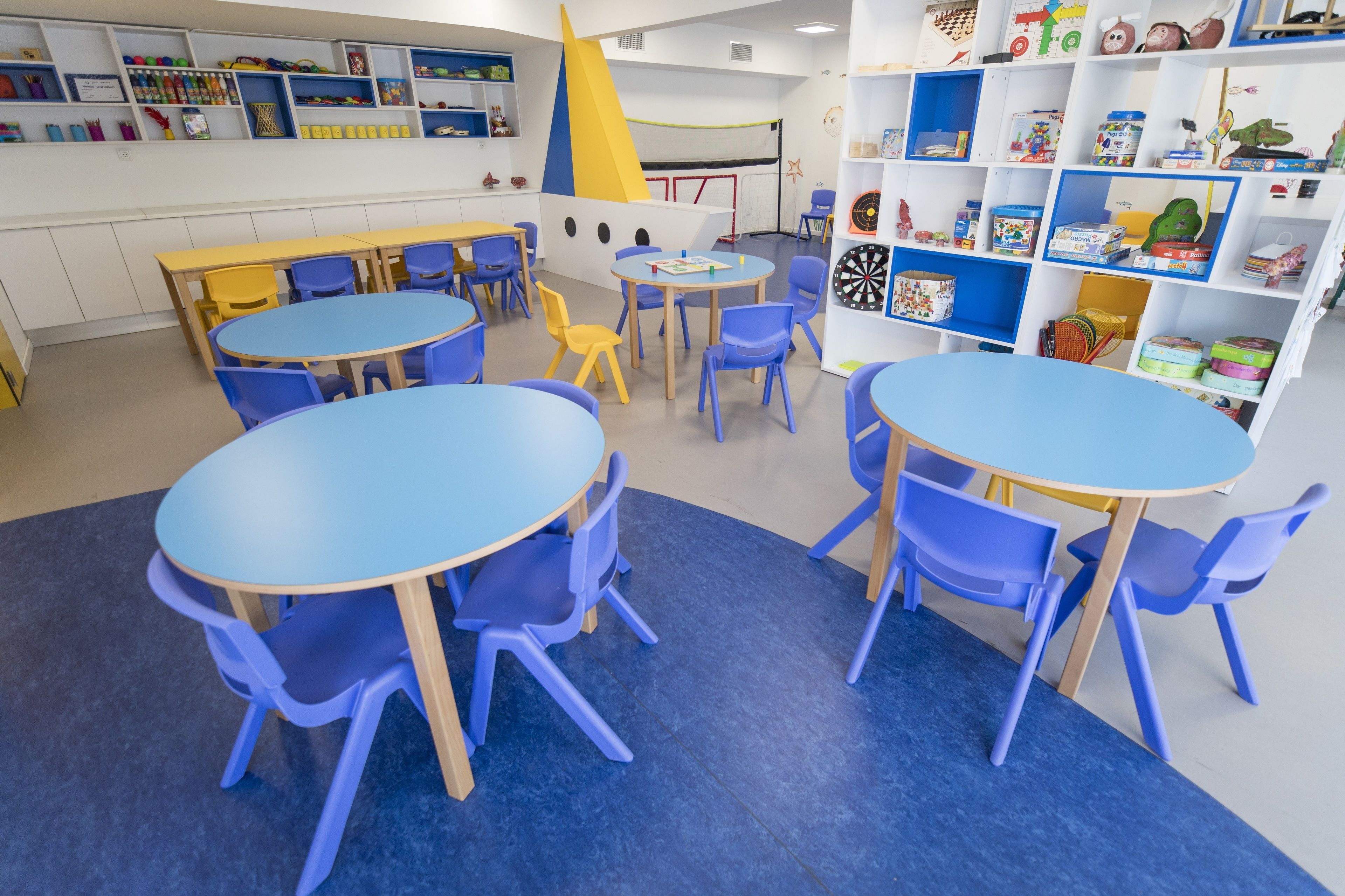 A children's playroom with round tables and blue chairs, shelves with toys, and educational materials.
