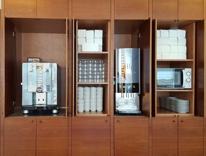 A kitchen with a water dispenser, stacked mugs, a coffee machine, a microwave, and a stack of plates. - Hotel Cesar Augustus (Cambrils)
