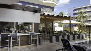 An outdoor seating area with tables and chairs, a bar with visible bottles, and a view of a building with balconies. - Hotel Cesar Augustus (Cambrils)