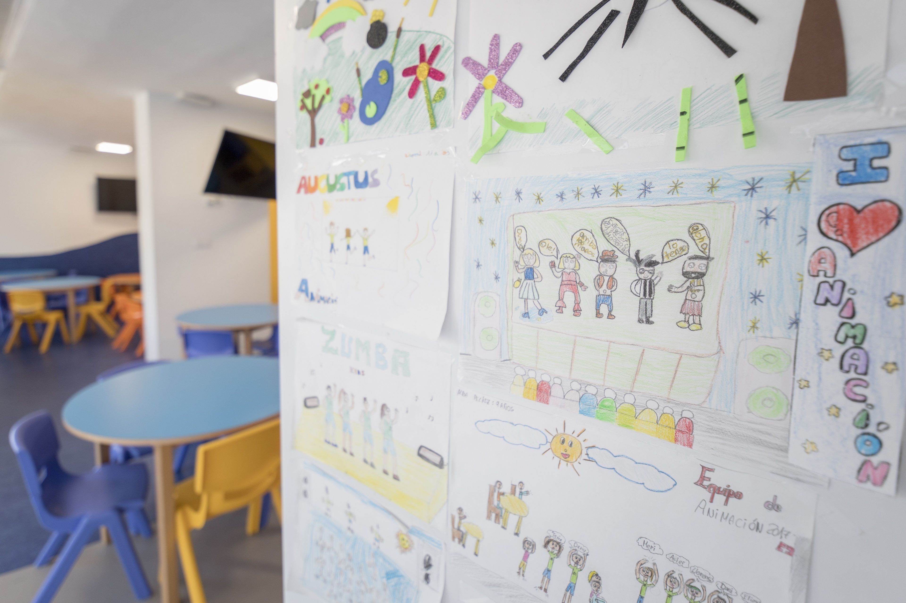 Een klaslokaal met kindertekeningen op de muur, tafels en stoelen.