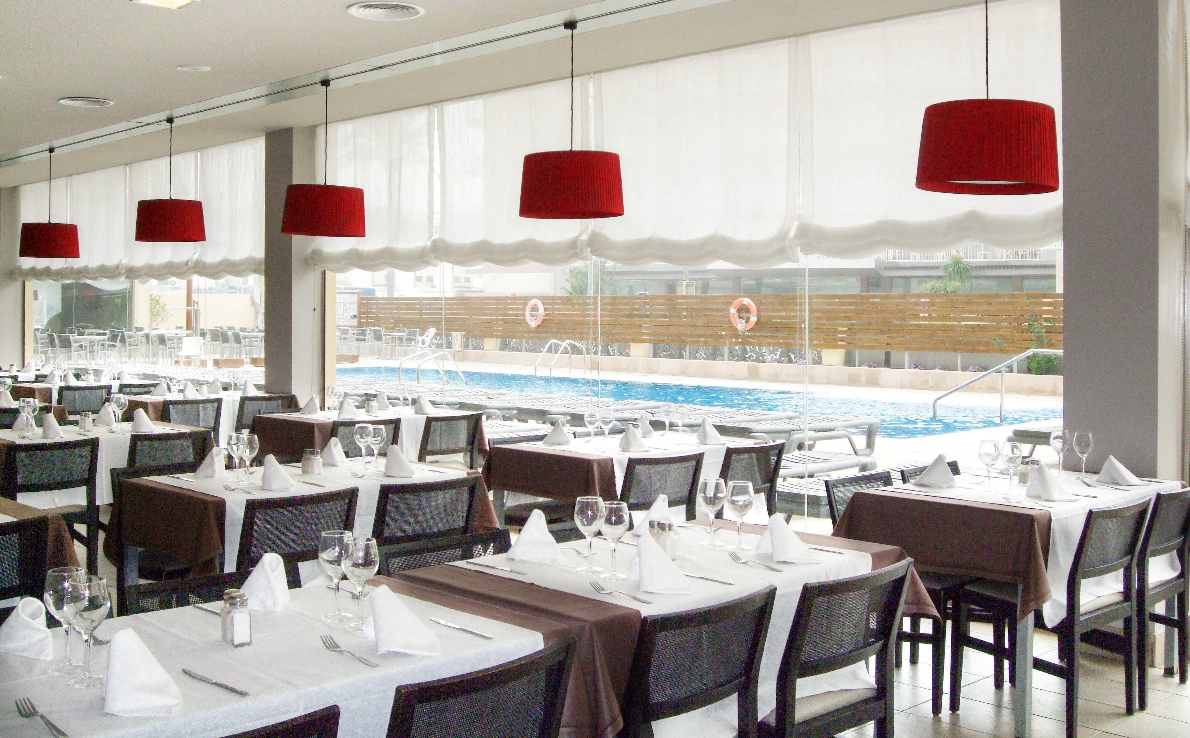 Un espace repas avec des tables dressées pour les invités, une piscine visible à travers les fenêtres et des lampes suspendues rouges.