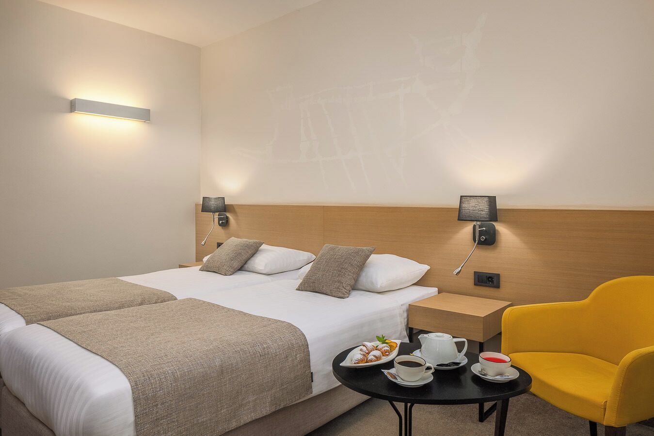 A hotel room with a double bed, two bedside tables, a small round table with breakfast, and a yellow armchair.