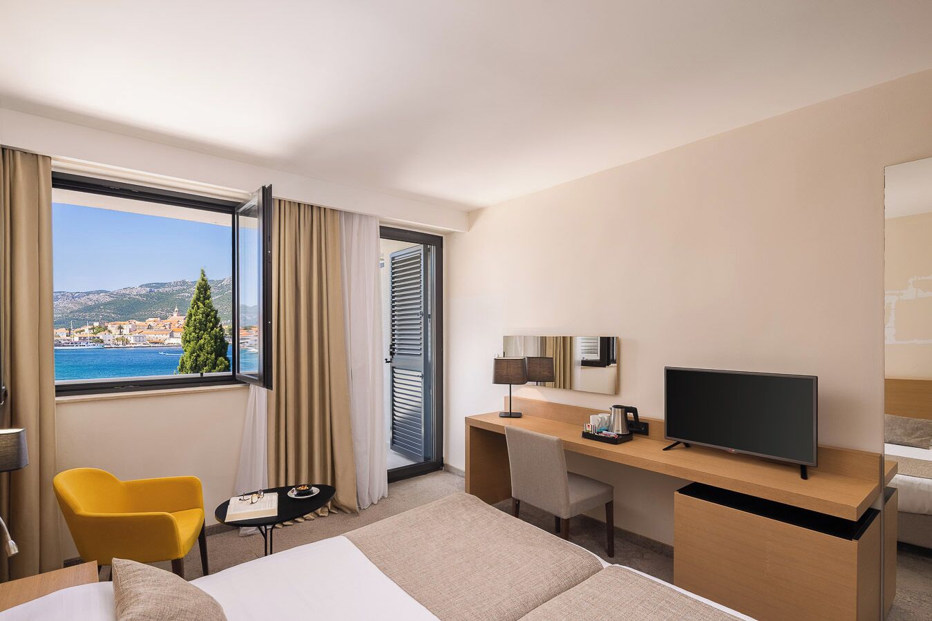 A hotel room with a bed, a desk, a chair, a TV, and a window with a view of a coastal town.