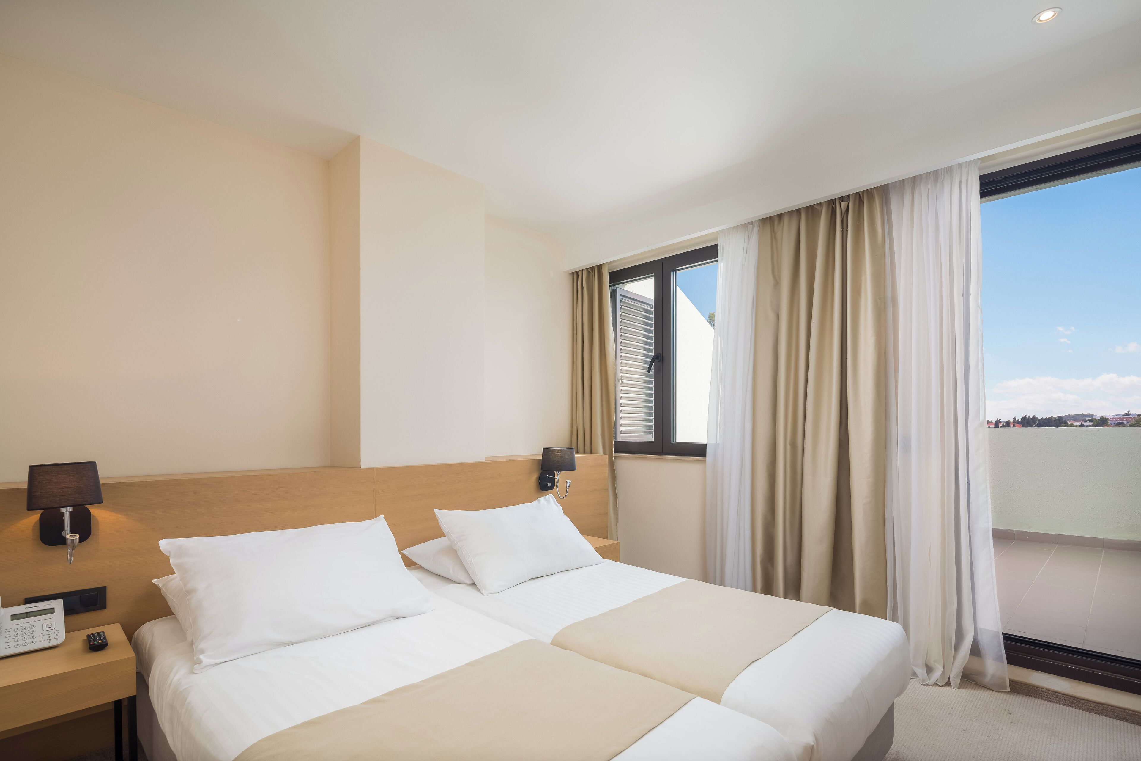 A hotel room with two beds, a bedside table with a lamp, a phone, and a window with curtains.