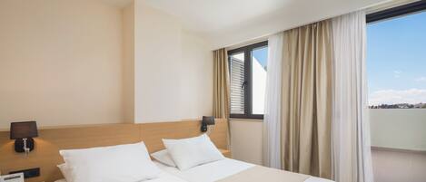 A hotel room with two beds, a bedside table with a lamp, a phone, and a window with curtains.