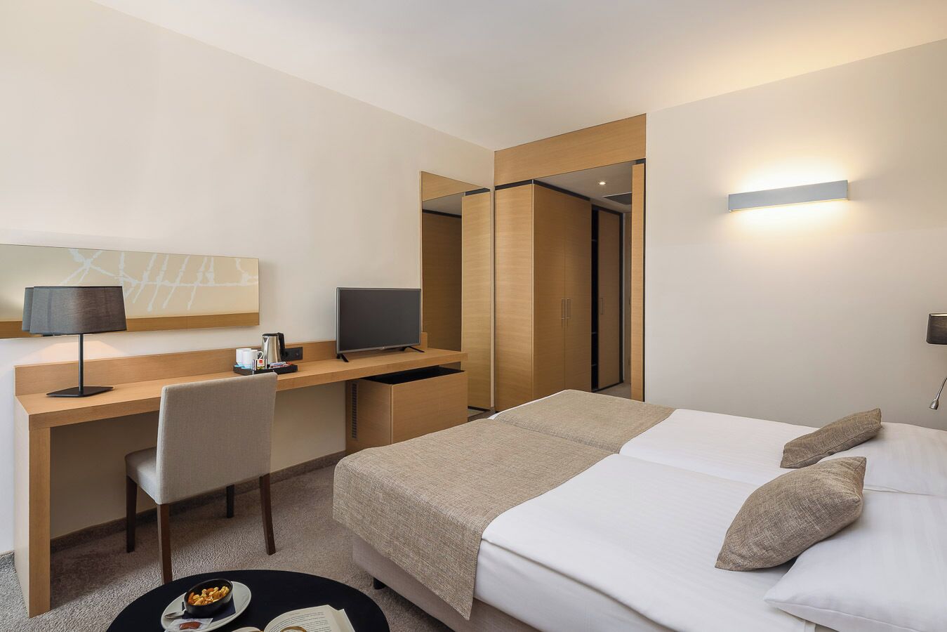 A hotel room with a bed, a desk with a computer, a chair, and a small table with a bowl of snacks.