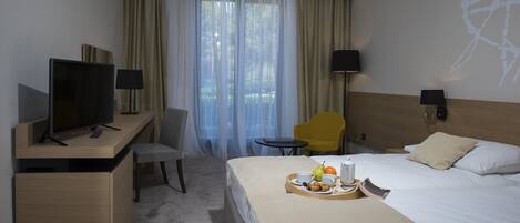 A hotel room with a bed, a desk with a TV, a chair, a small table, and a tray with breakfast items.
