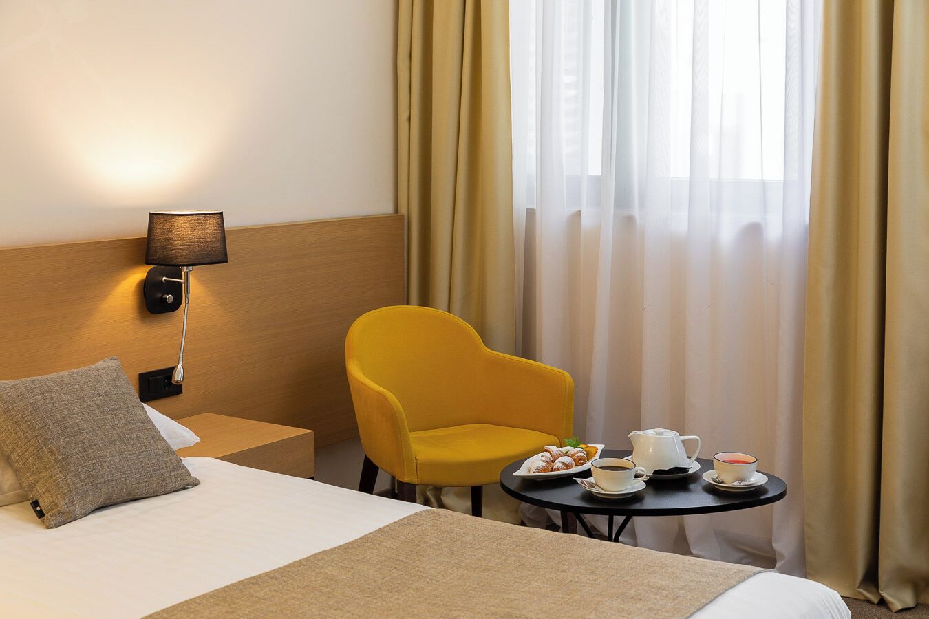 A hotel room with a bed, a yellow armchair, a small table with breakfast items, and a lamp.