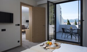A modern hotel room with a balcony, a flat-screen TV, a sofa, and a dining area with a table and chairs.
