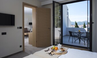 A modern hotel room with a balcony, a flat-screen TV, a sofa, and a dining area with a table and chairs.