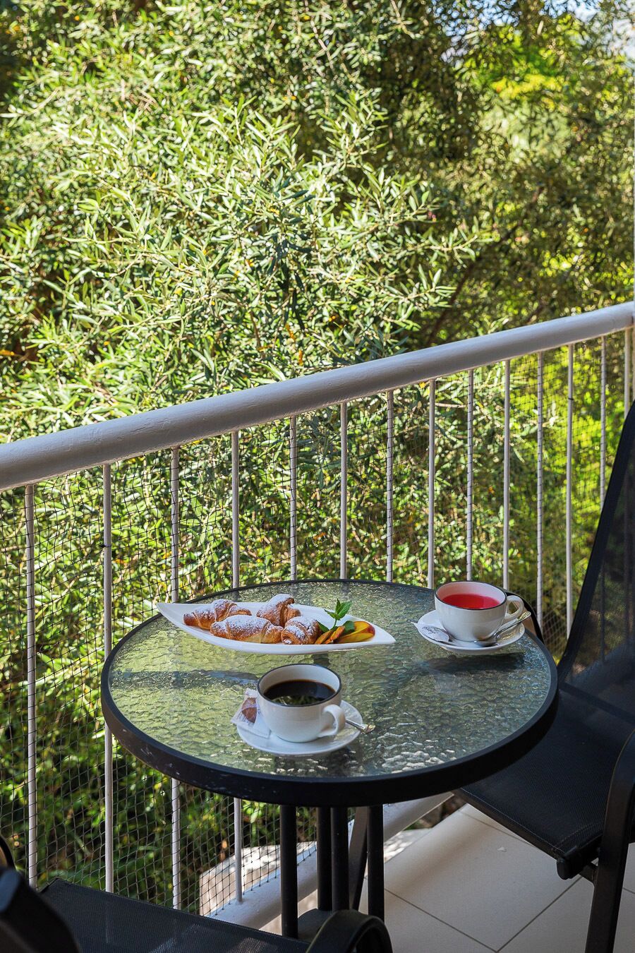 A round glass table with a white plate of pastries and a cup of coffee on a balcony.