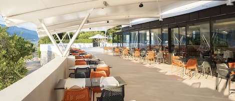 An outdoor terrace with tables and chairs, overlooking a body of water and mountains.