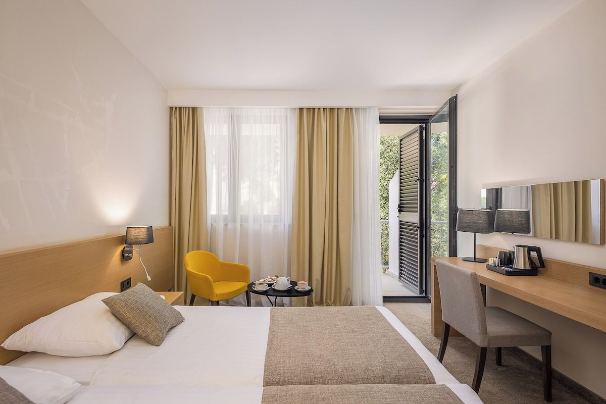 A hotel room with a bed, a desk, a chair, a small table, and a window with curtains.