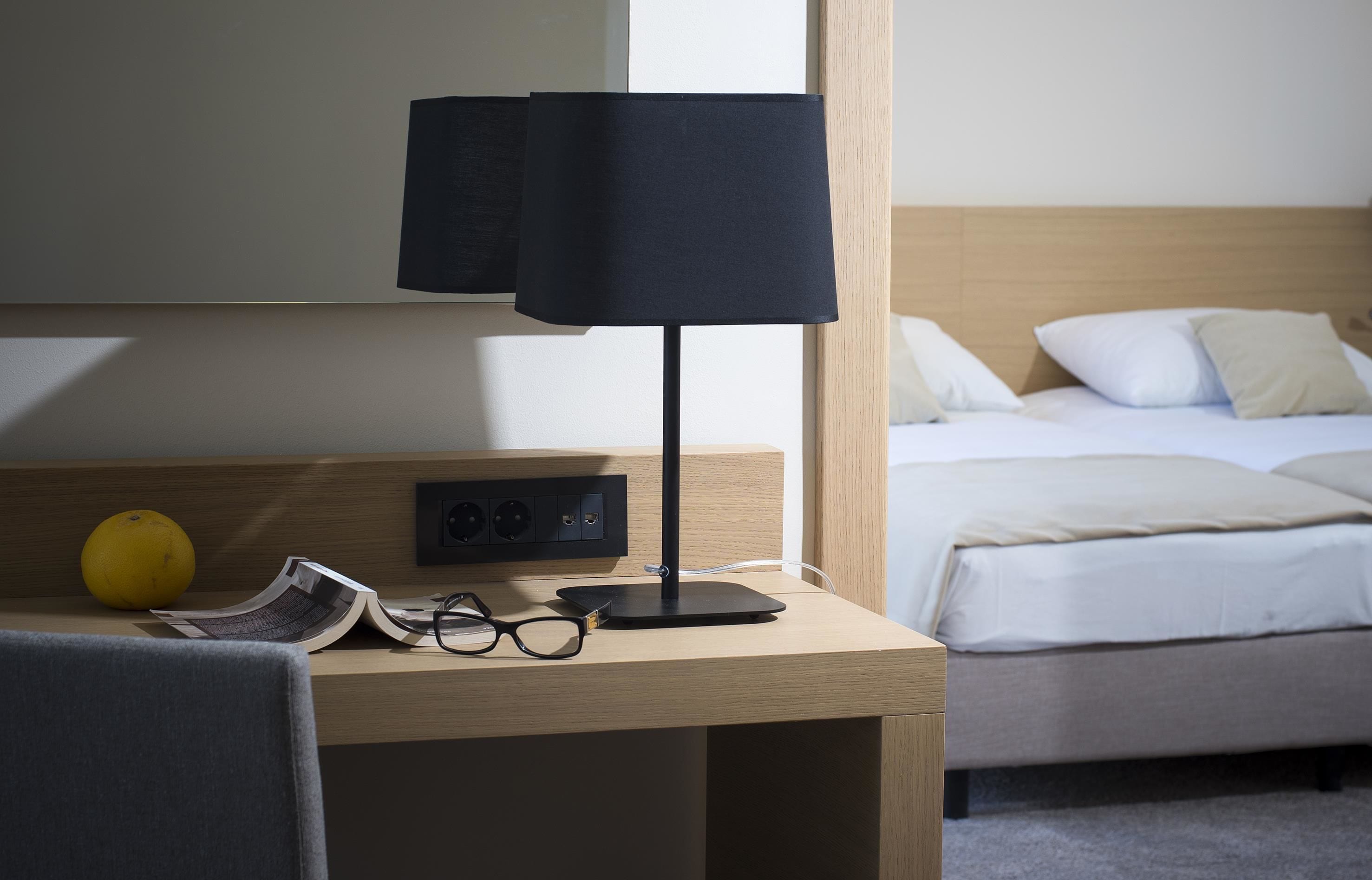 A hotel room with a wooden desk, a black lamp, a pair of glasses, and a yellow fruit.
