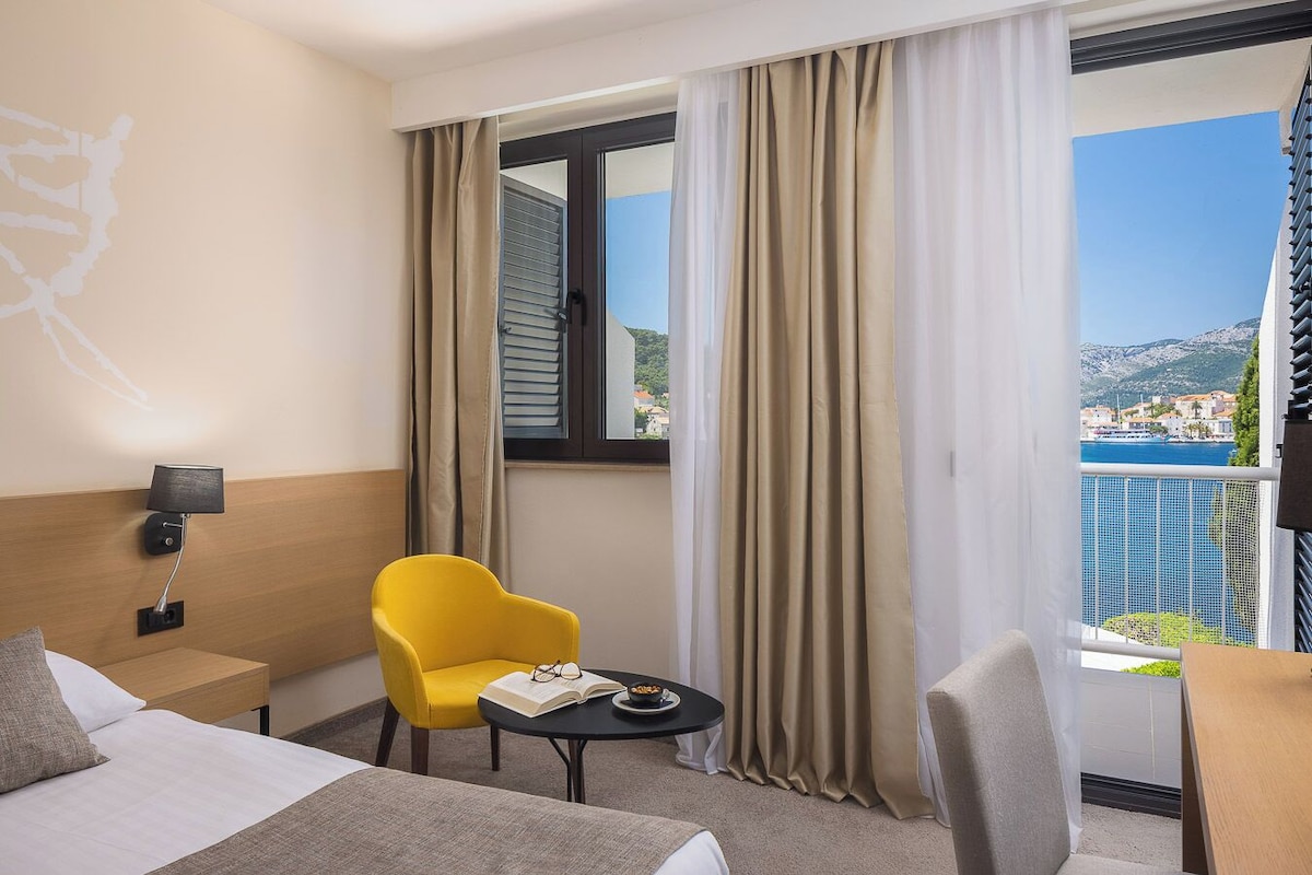 A hotel room with a bed, a yellow armchair, a small table with a book and a plate of nuts, a nightstand with a lamp, and a window with curtains.