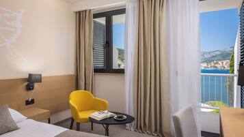 Une chambre d’hôtel avec un lit, un fauteuil jaune, une petite table avec un livre et une assiette de noix, une table de chevet avec une lampe et une fenêtre avec des rideaux.