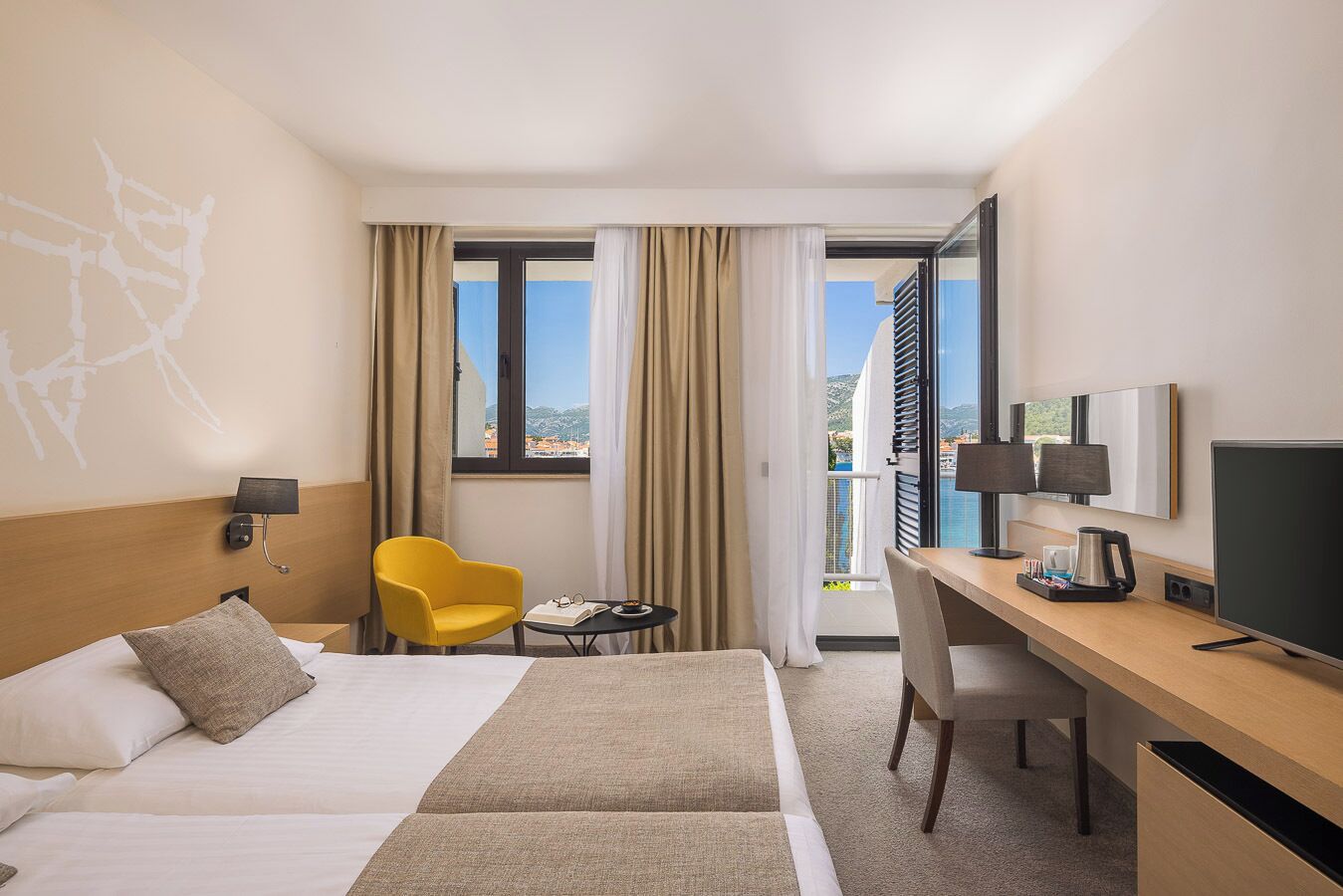 Quarto de hotel com cama, escrivaninha, cadeira, televisão e janela com cortinas.