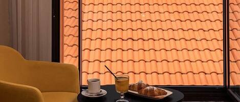 Hotellihuone, jossa on suuri ikkuna, josta avautuu näkymä rannikkokaupunkiin ja vuorille. Huoneessa on keltainen nojatuoli ja pieni pöytä, jonka päällä on juomaa ja leivonnaisia.