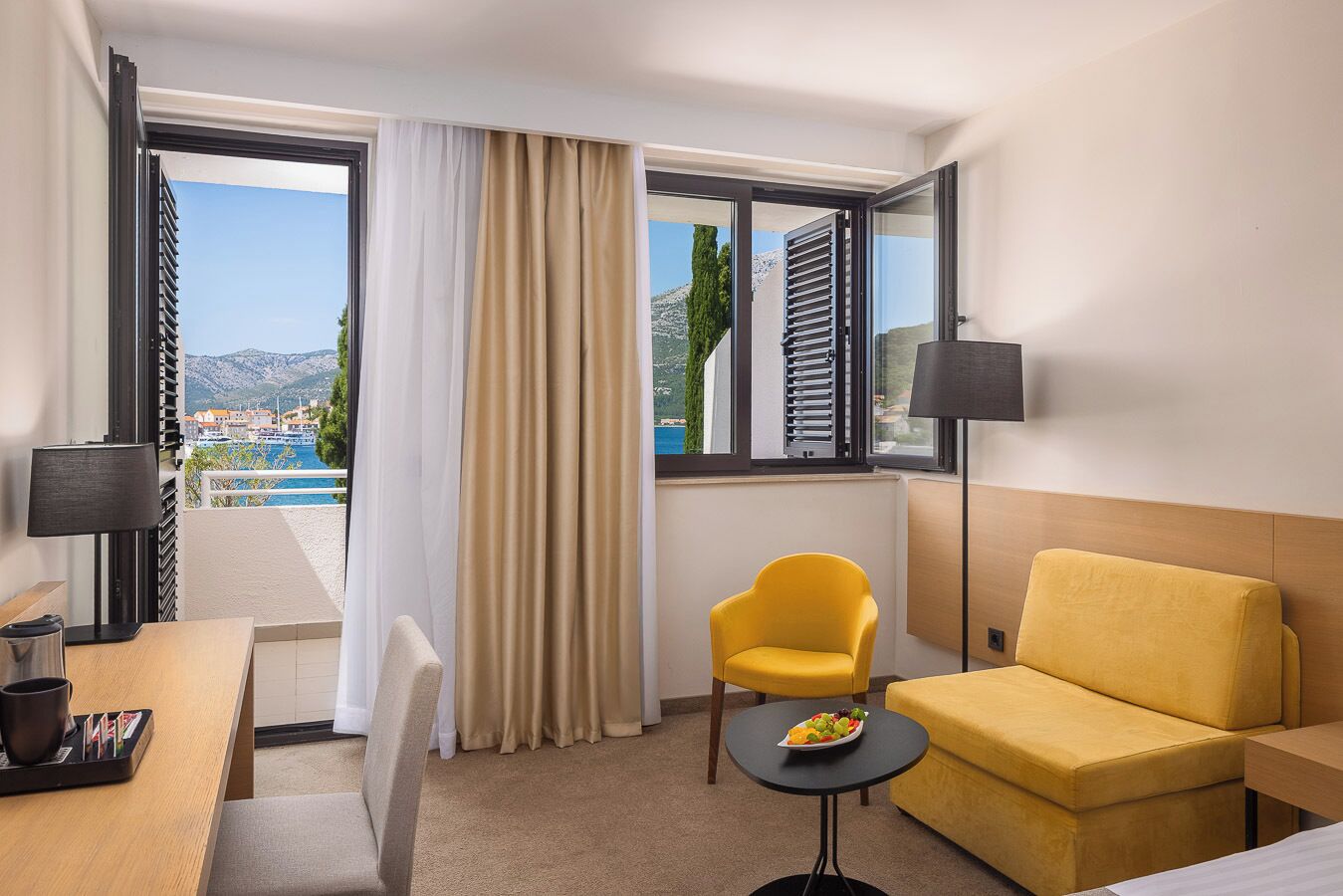 A hotel room with a balcony, a yellow armchair, a sofa, a small table with fruit, and a lamp.