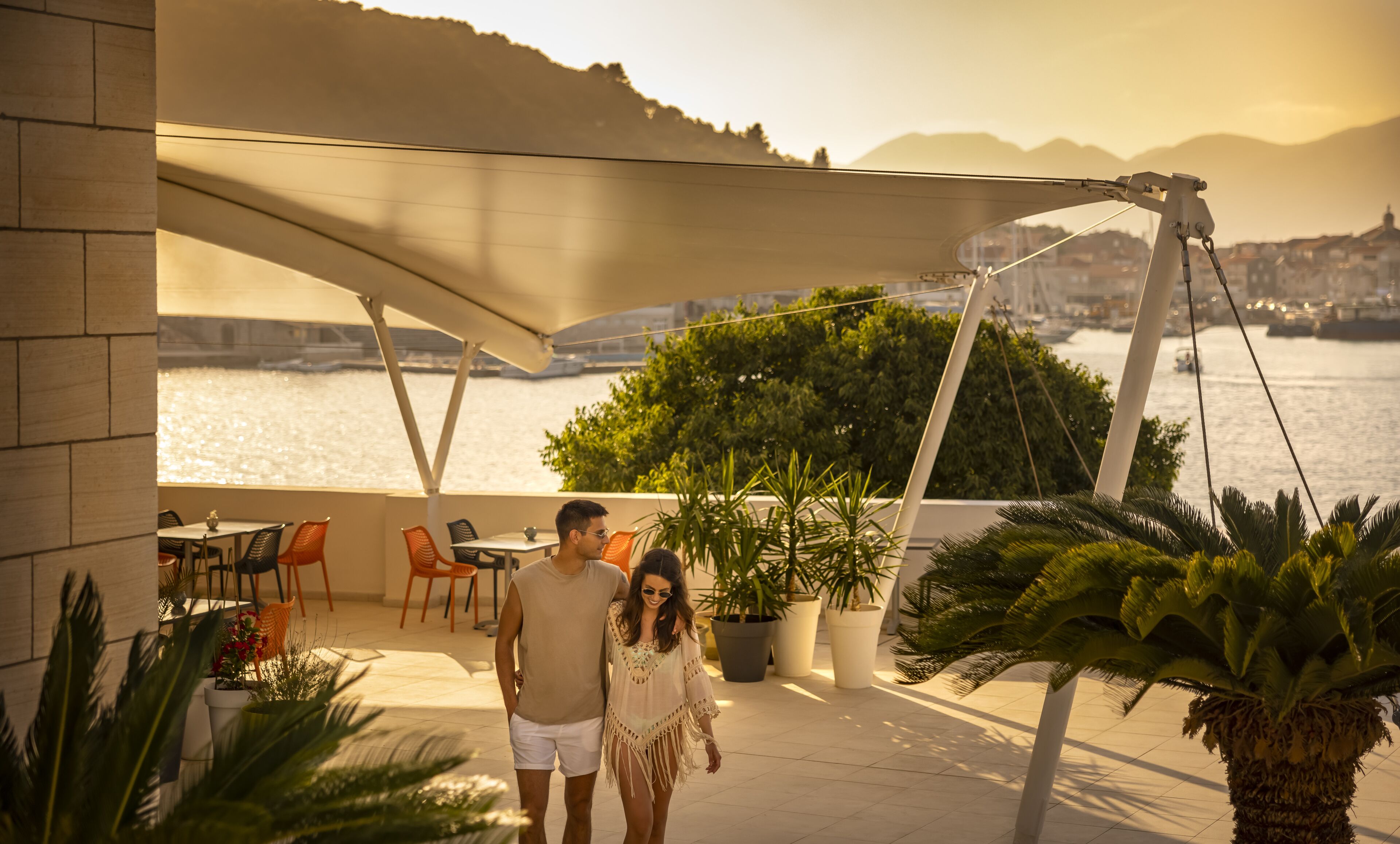 Una pareja caminando en una terraza con vista a un cuerpo de agua y montañas al fondo.