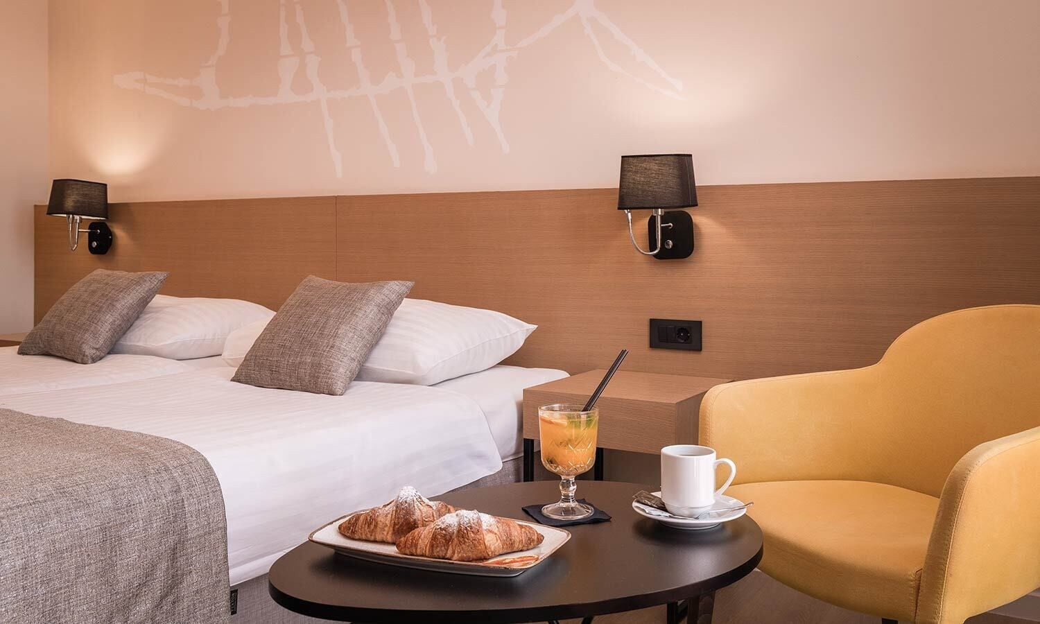 Et hotelværelse med en seng, to sengeborde med lamper, et bord med et bakværk, et glas juice og en gul lænestol.