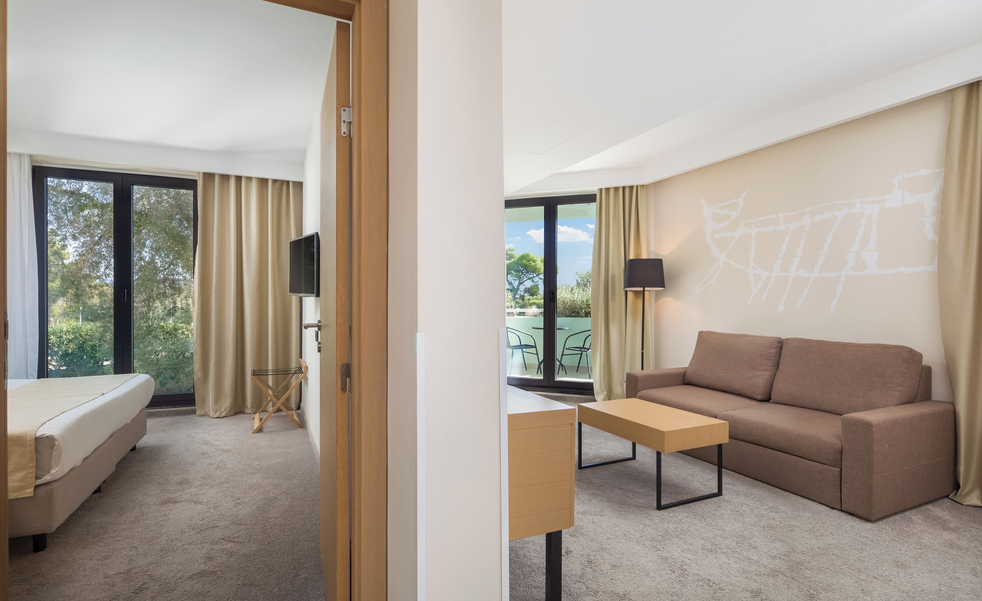 A hotel room with a bed, a desk, a sofa, and a balcony with a view of trees and a building.