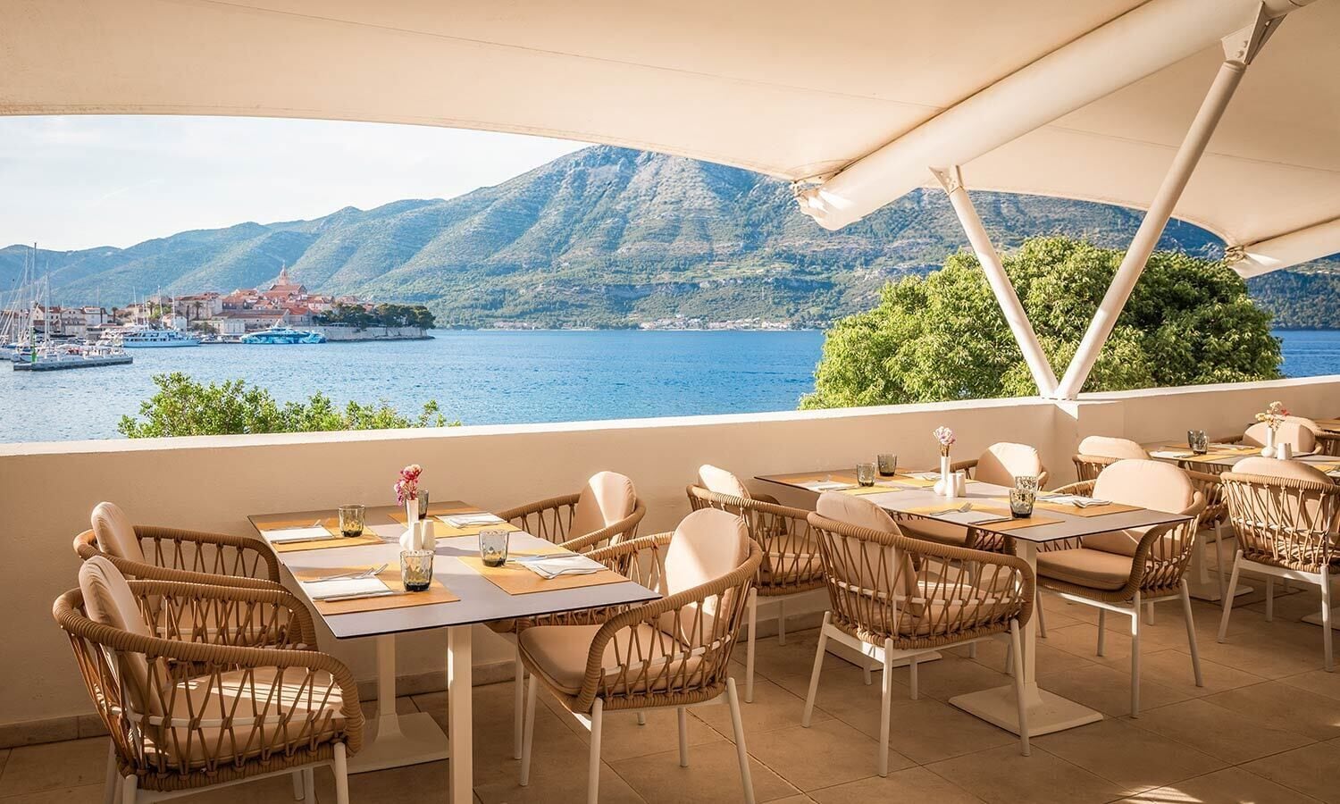 An outdoor dining area with wicker chairs and tables, overlooking a body of water with boats and a mountainous landscape in the background.