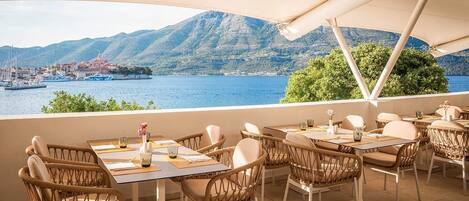 An outdoor dining area with wicker chairs and tables, overlooking a body of water with boats and a mountainous landscape in the background.
