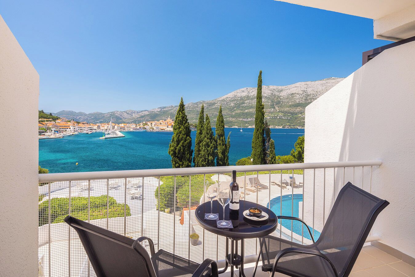 A balcony with a table and chairs, overlooking a coastal town and the sea.