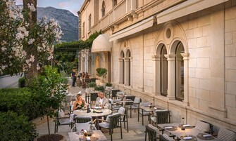 Um café ao ar livre com mesas e cadeiras, um edifício com janelas em arco e montanhas ao fundo.