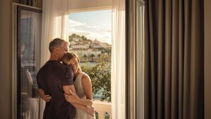 Una pareja se abraza en una habitación con un amplio ventanal que ofrece vistas panorámicas de edificios y vegetación.