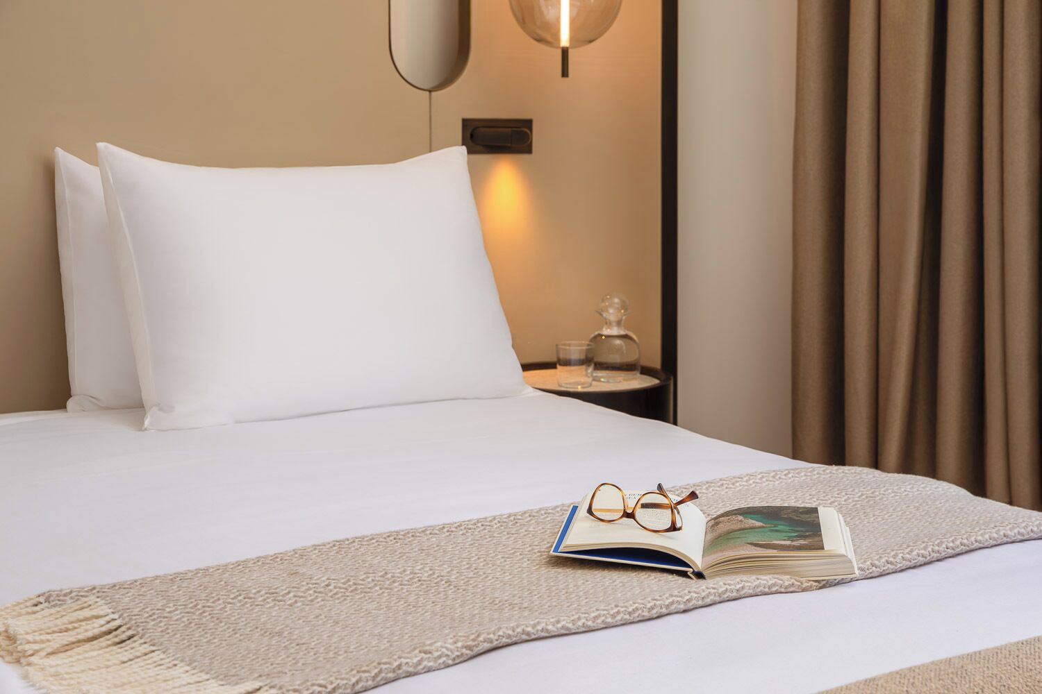 A neatly made bed with white linens, a pair of glasses, and an open book on a beige textured throw.